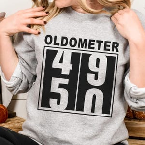 May include: A gray sweatshirt with the word "OLDOMETER" above a black and white graphic displaying the numbers "49" over "50". The sweatshirt is a casual style.