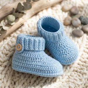 Peut inclure: Une paire de chaussons en crochet bleu clair dotés d'un revers côtelé et d'un petit bouton en bois sur le côté de chaque chaussure. Les chaussons sont réalisés avec un point de tricot dense, créant une surface texturée au bout arrondi.