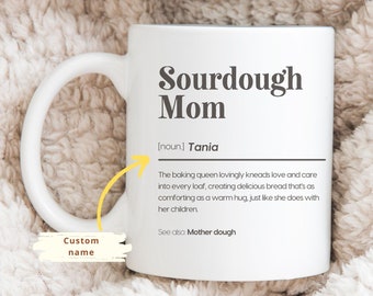 Puede incluir: Taza de cerámica blanca con el texto "Sourdough Mom" en negrita y oscura. La taza incluye una definición de "Sourdough Mom" y un espacio para un nombre personalizado. La taza está sobre una manta beige suave.