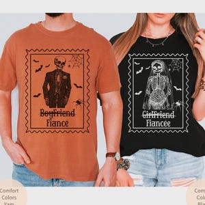 May include: Two matching Halloween-themed t-shirts. One is orange with a skeleton in a suit and the text "Boyfriend Fiance". The other is black with a skeleton bride and the text "Girlfriend Fiancee". Both shirts have a stamp-like design.