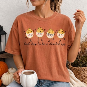 May include: A rust-colored short-sleeve t-shirt featuring a graphic of four cartoon deviled eggs with smiling faces and arms raised. The text below the eggs reads, bad day to be a deviled egg. One egg wears glasses. The shirt has a crew neckline.
