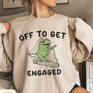 May include: Sand-colored sweatshirt with the text "OFF TO GET ENGAGED." The graphic features a green frog wearing a veil and riding a scooter. The sweatshirt is a long-sleeved crew neck.