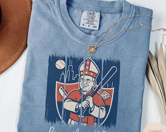 Könnte beinhalten: Blaues Comfort Colors T-Shirt mit einer Grafik eines Baseball spielenden Papstes mit einem Schläger. Das Design umfasst einen Baseball, eine Stadtlandschaft und den Text "Pope Leo XIV". Eine goldene Halskette liegt auf dem T-Shirt.