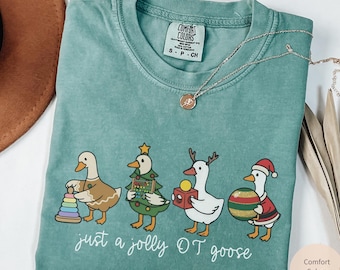 May include: Light green Comfort Colors t-shirt with a graphic of four ducks dressed for Christmas, holding toys and gifts. The text "just a jolly OT goose" is below the ducks. A silver necklace is on the shirt.