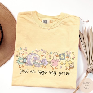 May include: Butter-colored Comfort Colors t-shirt with a whimsical graphic of geese dressed as medical professionals. The design includes the text "just an eggs-ray goose" and Easter-themed elements. A star-shaped necklace is visible.