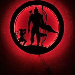 May include: A circular wall-mounted light fixture featuring a black silhouette of a standing archer with a quiver and bow, accompanied by a smaller kneeling figure. The design is illuminated from behind with a vibrant red glow.