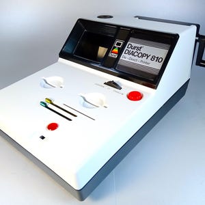 Peut inclure: Une imprimante Durst Diacopy 810 Dia-Direct-Printer vintage, une machine blanche et noire avec une poignée et des boutons rouges. La machine a une section supérieure noire et une base blanche, avec le logo Durst et le nom du modèle.
