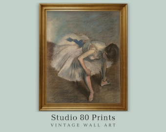 Könnte beinhalten: Gerahmter Vintage-Kunstdruck einer Ballerina, die ihren Schuh bindet. Das Kunstwerk in Pastelltönen zeigt eine Ballerina in einem weißen Tutu mit blauen Akzenten. Der goldfarbene Rahmen umgibt das Bild. Der Text "Studio 80 Prints" ist sichtbar.