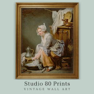 Puede incluir: Una impresión artística de pared vintage enmarcada que representa a una mujer lavando ropa. La obra de arte muestra a una mujer con gorro blanco y delantal, sentada cerca de una palangana. Una jarra de metal y un armario de madera son visibles en el fondo. El texto "Studio 80 Prints" está en la parte inferior.