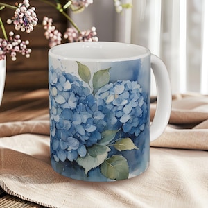 Op de afbeelding: Witte keramische mok met een aquarelontwerp van blauwe hortensia's en groene bladeren. De mok heeft een wit handvat en staat tegen een zachte, neutrale achtergrond, wat een bloemig thema suggereert.
