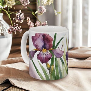 May include: White ceramic mug with a watercolour illustration of a purple iris flower with yellow and green accents. The floral design wraps around the mug, showcasing the flower's petals and leaves. The mug has a curved handle and is set against a neutral background.