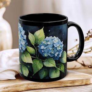 Op de afbeelding: Een zwarte keramische mok met een aquarelillustratie van blauwe hortensiabloemen met groene bladeren tegen een donkere, sfeervolle achtergrond. De mok heeft een glanzende afwerking, een gebogen handvat en een cilindrische vorm.