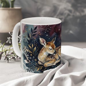 May include: A white ceramic mug featuring a watercolour illustration of a fawn resting among dark blue and purple foliage with gold accents. The design includes stylised leaves, branches, and small stars against a deep, moody background.