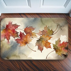 May include: A rectangular doormat featuring a watercolour illustration of several maple leaves in shades of red, orange, and green. The leaves are scattered across a soft, blended background of muted grey, beige, and earthy green tones.