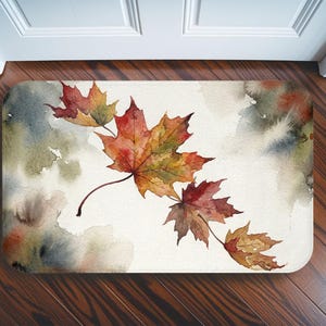 May include: A rectangular floor mat featuring a watercolour illustration of several maple leaves in shades of red, orange, and yellow. The leaves are arranged diagonally across a cream-coloured background with soft, blended washes of grey and green.