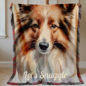 Puede incluir: Una manta tejida con un retrato detallado de un perro en tonos marrón, beige y blanco. La manta tiene un borde con flecos con detalles en rojo y las palabras "Let's Snuggle" en una fuente cursiva.