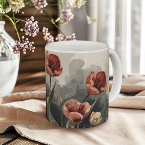 May include: White ceramic mug featuring a watercolor design of red and peach-colored tulips against a gray and beige background. The mug has a white handle and is set on a beige cloth.