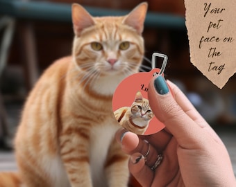 May include: A round, coral-colored pet tag with a silver-colored metal loop. The tag features a portrait of a ginger cat and the name "Lu". The tag is held in front of a blurred background with a ginger cat and the text "Your pet face on the tag".