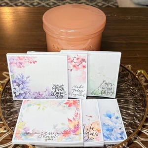 May include: A collection of floral-themed notepads with inspirational quotes, including "Start Fresh Dream Big" and "Jesus loves you." The notepads are arranged on a decorative tray, with a peach-colored jar in the background.