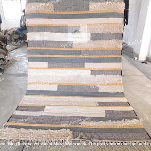 May include: A handwoven rug with a geometric pattern in shades of white, gray, and beige. The rug has a fringe border and is made of natural fibers.