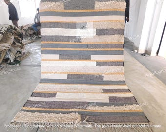 May include: A handwoven rug with a geometric pattern in shades of white, gray, and beige. The rug has a fringe border and is made of natural fibers.