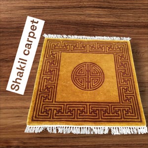 May include: A square, gold-coloured rug with a brown geometric pattern and a large, brown circle in the centre. The rug has a white fringe border.