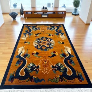 May include: An ornate, rectangular rug with a golden-brown field and a navy blue border. The rug features two large, stylized blue dragons facing each other, with a central circular design and cloud motifs. The rug has a fringed edge.