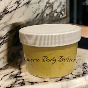 Może przedstawiać: Przezroczysty plastikowy słoik z białą pokrywką, wypełniony żółtym masłem do ciała. Tekst "Tumeric Body Butter" jest wydrukowany na słoiku kursywą. Słoik stoi na marmurowym tle.