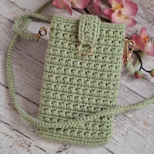 May include: A sage green crochet crossbody bag featuring a textured stitch pattern, a vertical flap closure with a gold-toned metal ring, and a matching crocheted strap attached with gold-toned swivel clasps on both sides of the rectangular pouch.