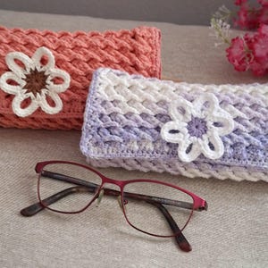 Peut inclure: Deux étuis à lunettes crochetés, un corail et un lavande, chacun avec une application de fleur. Une paire de lunettes à monture rouge repose devant les étuis. Les étuis ont un aspect texturé et tissé.