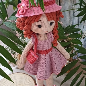 Puede incluir: Una muñeca de crochet que lleva un vestido rosa, un sombrero rosa con una flor y zapatos rojos. La muñeca tiene el pelo castaño y los ojos marrones.