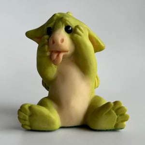 Puede incluir: Una figura caprichosa de una criatura verde con vientre y cara de color beige claro. La criatura tiene grandes ojos negros, una nariz rosada y saca la lengua. Está sentada con las manos cubriendo sus ojos y las orejas levantadas.