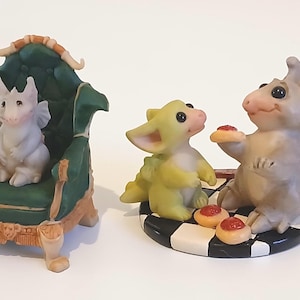 Puede incluir: Tres pequeñas figuritas de dragones, una sentada en un trono verde, otra sentada en una base de tablero de ajedrez blanco y negro, y la última sentada en una base de tablero de ajedrez blanco y negro con una rebanada de pizza roja en la boca.