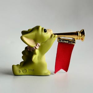 May include: A light green figurine of a dragon playing a gold trumpet with a red banner. The dragon has a pink bow and the year 2001 is printed on the base. The figurine is on a white background.