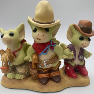 May include: Three green ceramic figurines of dragons wearing cowboy hats and boots. The dragon on the left is holding a brown horse figurine. The dragon in the middle is wearing a red bandana. The dragon on the right is wearing a purple vest.