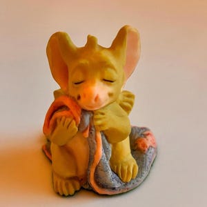 May include: A small, yellow figurine of a dragon-like creature, with closed eyes, holding a blanket. The blanket is orange and blue. The figurine is resting on a blue and pink patterned base.