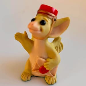 May include: A whimsical figurine of a yellow dragon wearing a red hat with a gold band. The dragon holds a cream-colored envelope with a red heart seal. The dragon has large black eyes and is gesturing with one arm.