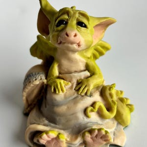 May include: A whimsical figurine of a small, green dragon with large ears and wings. The dragon has a sad expression and is seated on a cream-colored base. The figurine is painted with yellow, pink, and cream tones.