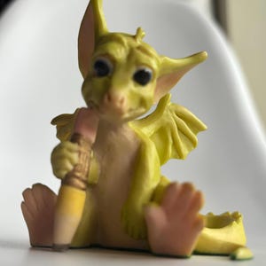 Puede incluir: Una figura caprichosa de un dragón amarillo sentado sosteniendo un lápiz. El dragón tiene ojos grandes y expresivos, alas pequeñas y una barriga y orejas de color rosa claro. El lápiz es amarillo con una goma de borrar rosa.