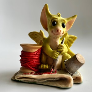 Puede incluir: Una figura caprichosa de un dragón amarillo con grandes orejas y alas, sosteniendo una aguja. Está sentado junto a una bobina de hilo rojo y un dedal. Un artículo decorativo encantador.