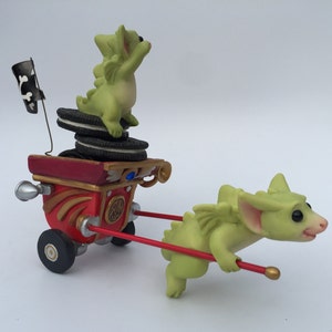 May include: A green dragon figurine pulling a red wagon with a green dragon figurine on top. The wagon has a pirate flag and a stack of chocolate chip cookies.