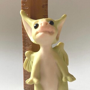 Puede incluir: Una pequeña figura de dragón verde y blanco con alas, de pie junto a una regla. El dragón mide aproximadamente 7,5 cm de alto.