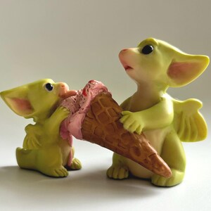 May include: Two small, yellow dragon figurines sharing an ice cream cone. One dragon is licking the pink ice cream, while the other holds the cone. The cone is brown, and the dragons have large ears and small wings.