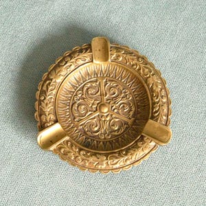 Könnte beinhalten: Ein runder, antiker Aschenbecher aus Messing mit einem aufwendigen, geprägten Design. Der Aschenbecher hat drei Zigarettenablagen und ein zentrales dekoratives Muster. Der Rand ist mit einem sich wiederholenden Blumenmotiv versehen.