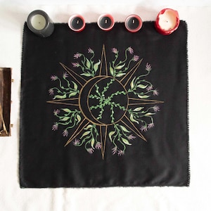Peut inclure: Nappe d'autel noire avec un motif de soleil et de lune dorés avec des accents floraux. Le motif est brodé de feuilles vertes et de fleurs roses. Le tissu a un bord effiloché. Plusieurs bougies sont placées autour du tissu.