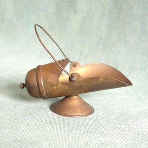 May include: A vintage brass sugar scuttle with a scoop-shaped bowl and a handle. The scuttle is mounted on a small, round base. The brass has a warm, aged patina, suggesting its antique nature. The handle is arched and attached to the bowl with small hinges.