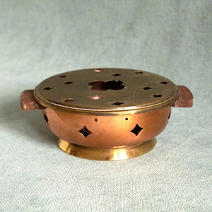May include: A copper-colored incense burner with a brass lid featuring decorative cutouts. The burner has wooden handles and diamond-shaped cutouts around the sides. The base is a lighter brass color.