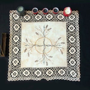 Peut inclure: Une nappe d'autel carrée de couleur crème avec une bordure en dentelle ajourée détaillée. Le centre présente un dessin dessiné à la main d'un cercle, d'une croix et d'éléments botaniques en marron et vert. Plusieurs bougies de tailles et de couleurs variées sont disposées autour du tissu.
