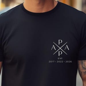 May include: Black crew neck t-shirt with a white logo on the upper left chest. The logo features crossed lines with the letters "P", "A", and "X", and the text "EST 2017-2022-2026" below it. The shirt is made of a soft material.
