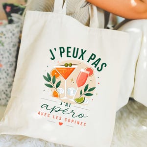 Peut inclure: Sac fourre-tout en toile écrue avec un motif de cocktail coloré. Le sac porte l'inscription "J' PEUX PAS J'AI apéro AVEC LES COPINES" en vert et rouge, ainsi que des illustrations de cocktails, de fruits et de glaçons.